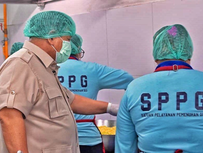 People in hairnets and face masks work at a food preparation station a supervisor in a tan uniform observes two staff wearing blue sppg shirts beside a metal prep area